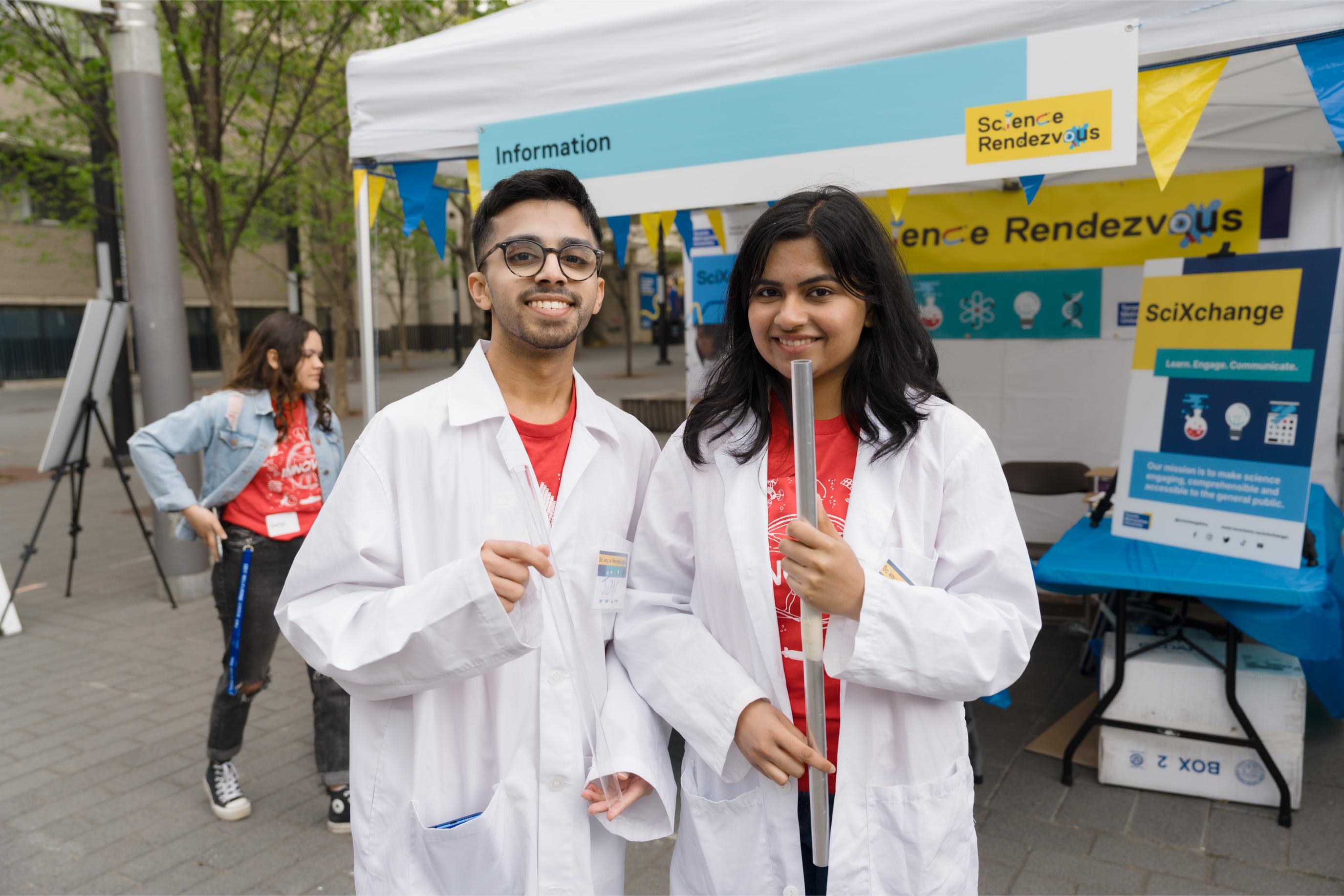 Two pocket science volunteers in white lab coats holding two clear cylinders are posing and smiling at the camera.